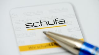 Close-up of a Schufa document featuring the Schufa logo in black and yellow. In the foreground, the tip of a blue ballpoint pen is blurred over the paper.