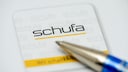 Close-up of a Schufa document featuring the Schufa logo in black and yellow. In the foreground, the tip of a blue ballpoint pen is blurred over the paper.