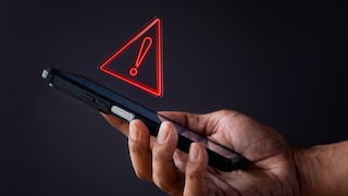 A hand holds a black smartphone against a dark background. Above the device hovers a red warning symbol in the shape of a glowing triangle with an exclamation mark. The image represents a security warning or potential risk associated with smartphones.