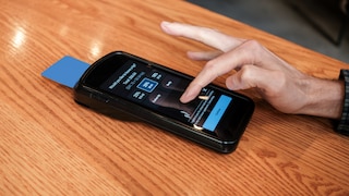 A card reader sits on a light wooden table. The display shows several tip options in percentages, including 18, 20, and 25 percent, along with a button for a custom amount. A hand taps the screen. A blue payment card is inserted into the side of the terminal.