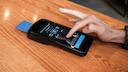 A card reader sits on a light wooden table. The display shows several tip options in percentages, including 18, 20, and 25 percent, along with a button for a custom amount. A hand taps the screen. A blue payment card is inserted into the side of the terminal.