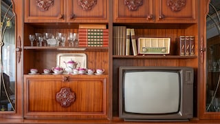 An old tube TV sits in a dark wooden wall unit. Next to it are books, glasses, a tea set, and a radio.