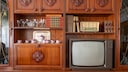 An old tube TV sits in a dark wooden wall unit. Next to it are books, glasses, a tea set, and a radio.
