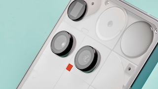 The back of a white Nothing smartphone is shown at an angle. Visible are three large round camera modules, a small sensor, and the characteristic transparent design with geometric elements beneath the back cover, typical of Nothing. The device is set against a light turquoise background.