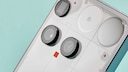 The back of a white Nothing smartphone is shown at an angle. Visible are three large round camera modules, a small sensor, and the characteristic transparent design with geometric elements beneath the back cover, typical of Nothing. The device is set against a light turquoise background.