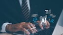 A man in a suit sits at a desk, working with AI on his laptop.