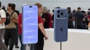Two blue Apple smartphones are displayed on metal stands in a brightly lit showroom. On the left, the front with the screen turned on is visible, while on the right, the back with three camera lenses is shown. In the blurred background, several people and additional display areas can be seen.