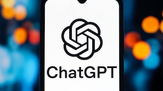 A smartphone is positioned upright against a blurred background featuring blue, orange, and red light spots. The display shows the black ChatGPT logo on a white background, with the word "ChatGPT" written beneath it. At the top edge of the display, there's a small notch for the front camera. The smartphone occupies most of the image and is centrally placed.