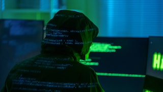 A man in a hoodie—allegedly a hacker—sits in front of a computer, surrounded by a flurry of data code.