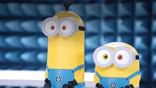 Two yellow routers designed like Minions stand side by side on a white surface. The larger model on the left features a longer body, gray goggles with two large eyes, three black strands of hair on its head, and blue overalls. The smaller model on the right also has gray goggles, two differently colored eyes, and blue overalls. Both devices have black hands and feet. In the background, a blurred blue structure with rectangular shapes is visible.