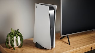 A white PlayStation 5 stands upright on a wooden TV stand next to a flat-screen TV. To the left, there's a potted plant with trailing green leaves. The gaming console faces forward, with ports and the disc drive visible on the side. The living room is simply furnished and softly lit.