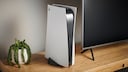 A white PlayStation 5 stands upright on a wooden TV stand next to a flat-screen TV. To the left, there's a potted plant with trailing green leaves. The gaming console faces forward, with ports and the disc drive visible on the side. The living room is simply furnished and softly lit.