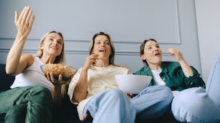 Three people are sitting on a sofa, looking at a screen off-camera. One person is holding a bowl of popcorn, while the other two have snack bowls. The shot captures the group from a low angle in a bright living room.