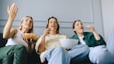 Three people are sitting on a sofa, looking at a screen off-camera. One person is holding a bowl of popcorn, while the other two have snack bowls. The shot captures the group from a low angle in a bright living room.