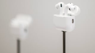 White Apple AirPods Pro in an open charging case on a slim black stand. In the background, another AirPods case is visible, though blurred. The photo is brightly lit, showcasing the headphones against a gray background with shallow depth of field.