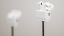 White Apple AirPods Pro in an open charging case on a slim black stand. In the background, another AirPods case is visible, though blurred. The photo is brightly lit, showcasing the headphones against a gray background with shallow depth of field.