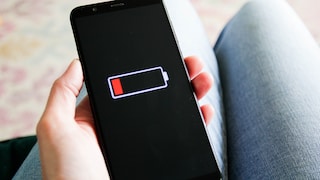 A person holds a smartphone in their hand. The black display shows an almost empty battery with a red charging indicator. In the background, the person's legs are visible but out of focus.
