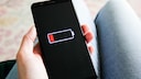 A person holds a smartphone in their hand. The black display shows an almost empty battery with a red charging indicator. In the background, the person's legs are visible but out of focus.