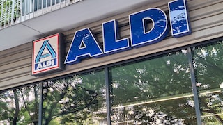 Exterior view of an Aldi store featuring a large blue Aldi sign and logo on the building facade. Below the sign are large windows reflecting trees and their leaves. Above the entrance, a balcony with a metal railing is visible.