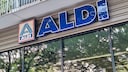 Exterior view of an Aldi store featuring a large blue Aldi sign and logo on the building facade. Below the sign are large windows reflecting trees and their leaves. Above the entrance, a balcony with a metal railing is visible.