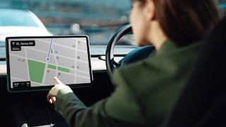 A person is sitting behind the wheel of a car, operating a large screen on the dashboard. The display shows a map view of a navigation app with route and street information. The steering wheel is located to the right of the screen, with blurred traffic visible in the background.