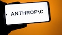A smartphone in landscape mode displays the Anthropic logo on a bright screen. The device is held against an orange background.