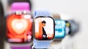 An Apple Watch with a light blue band is in the foreground. The display shows a watch face featuring a person in profile. In the blurred background, there are more Apple Watches with various bands and colorful displays.