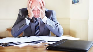 The image shows a person in a suit sitting at a desk, resting their head in both hands. Several documents and a closed black briefcase are on the desk. In the background, a bright office space with a framed picture on the wall is visible.