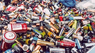 A large pile of used batteries of various sizes and brands is visible. The pile includes numerous AA, AAA, C, and D cells, along with other types of batteries in different colors, all jumbled together. Some batteries show signs of wear and corrosion. In the background, there are more batteries and plastic bags.