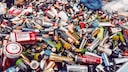 A large pile of used batteries of various sizes and brands is visible. The pile includes numerous AA, AAA, C, and D cells, along with other types of batteries in different colors, all jumbled together. Some batteries show signs of wear and corrosion. In the background, there are more batteries and plastic bags.
