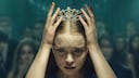 Close-up of a young person wearing a crown. Both hands are holding the crown firmly, while several people can be seen in the blurred background.