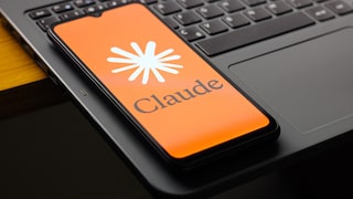 A smartphone with an orange Claude start screen is positioned at an angle on the keyboard of an open laptop. The display shows the Claude logo and the word "Claude." The close-up shot captures the device on a dark table.