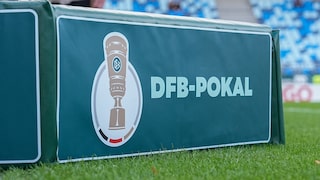 DFB-Pokal logo on a green advertising board at the edge of a soccer stadium field. In the background, blurred blue and white stadium seats and parts of the grass are visible.