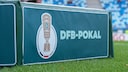 DFB-Pokal logo on a green advertising board at the edge of a soccer stadium field. In the background, blurred blue and white stadium seats and parts of the grass are visible.