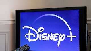 A television with the Disney+ logo on a blue background, with a hand holding a remote in front of the screen.
