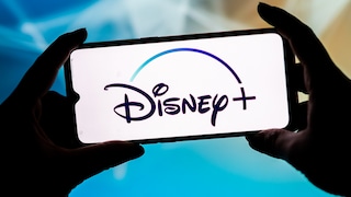 Two hands hold a smartphone in landscape mode. The bright display prominently features the Disney+ logo. In the blurred background, shades of blue, turquoise, and light beige dominate.