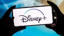 Two hands hold a smartphone in landscape mode. The bright display prominently features the Disney+ logo. In the blurred background, shades of blue, turquoise, and light beige dominate.