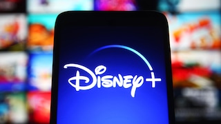 A screen displaying the Disney+ logo is in the foreground. In the background, several colorful preview images of movies and series are visible on a streaming interface, though they are blurred.