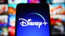 A screen displaying the Disney+ logo is in the foreground. In the background, several colorful preview images of movies and series are visible on a streaming interface, though they are blurred.