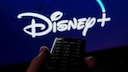 A remote control is held in front of a television. The Disney+ logo is visible, though blurred, on a blue background on the screen.