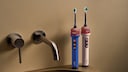 Two electric toothbrushes from Dyson sit on the edge of a sink next to a wall-mounted faucet. One toothbrush is blue, the other light pink. Both have a transparent section near the top, just below the brush head. A beige wall is visible in the background.