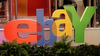 Large colorful eBay logo with red, blue, yellow, and green letters in front of a trade show booth. In the foreground, there are several plants, and in the background, glass walls and parts of the exhibition area are visible.