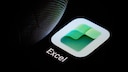 Close-up of a smartphone display featuring the Excel app. The green-and-white Excel logo is visible against a black background. To the left, a finger is either hovering over or touching the app. Below the icon, the word "Excel" is displayed.