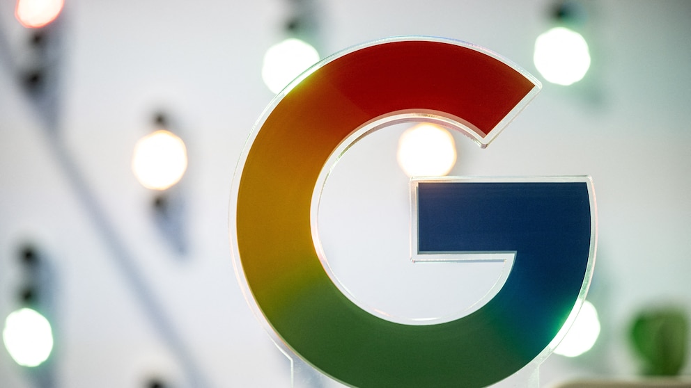 A large Google logo in the company's colors of red, yellow, green, and blue. The logo is positioned on a transparent stand in the foreground. In the blurred background, several bright light sources are visible in an indoor setting.