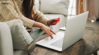 A person sits at a wooden table with a laptop and holds a red card in their hand. A smartphone is also on the table. In the background, a light-colored sofa and a window are visible.