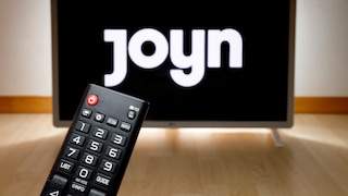 A remote control lies in front of a television. The logo of the streaming service Joyn is visible on the screen in the background.