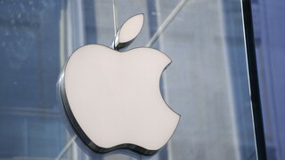 Close-up of a white Apple logo on a glass wall. In the background, blurred structures of a modern building in shades of blue and gray are visible.