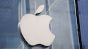 Close-up of a white Apple logo on a glass wall. In the background, blurred structures of a modern building in shades of blue and gray are visible.