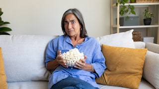 A person in a loose blue shirt sits on a light-colored sofa, holding a glass bowl of popcorn. In the background, there's a shelf with plants, storage boxes, and decorations. Several beige and mustard-colored pillows are on the sofa. The person is looking directly at the camera. The photo was taken in a bright living room.