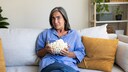 A person in a loose blue shirt sits on a light-colored sofa, holding a glass bowl of popcorn. In the background, there's a shelf with plants, storage boxes, and decorations. Several beige and mustard-colored pillows are on the sofa. The person is looking directly at the camera. The photo was taken in a bright living room.